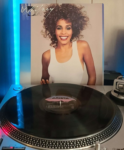 Image shows a turntable with a black vinyl record on the platter. Behind the turntable vinyl album outer sleeve is displayed. The front cover shows Whitney Houston in a white tank top looking at the camera and smiling brightly. 