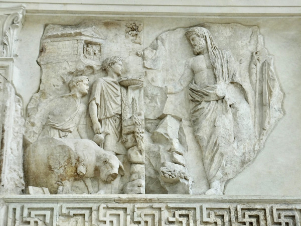 Relief panel showing two youthful figures wearing wreaths and accompanied by a cow approaching a beard man who is veiled as though for ritual purpose. Another figure stands behind the man but only a hand holding a staff can be seen.
