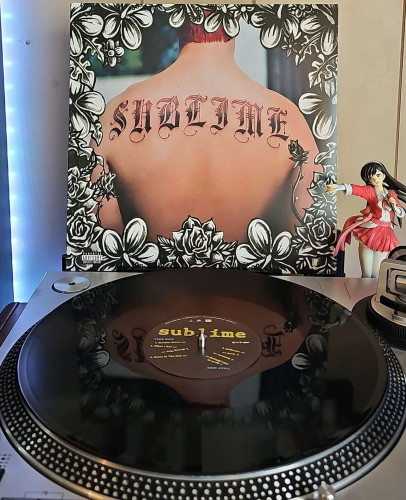 A black vinyl record sits on a turntable. Behind the turntable, a vinyl album outer sleeve is displayed. The front cover shows the back of a person with a SUBLIME tattoo. 

To the right of the album cover is an anime figure of Yuki Morikawa singing in to a microphone and holding her arm out. 