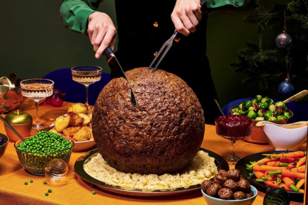 Meatball that is larger than a basketball,  sitting on a platform of noodles and being carved by someone wearing fancy gear and green felt shirt. Pile of peas and potatoes and other food is on the gable next to the incredibly large meatball. Gravy and cranberries nearby as well.