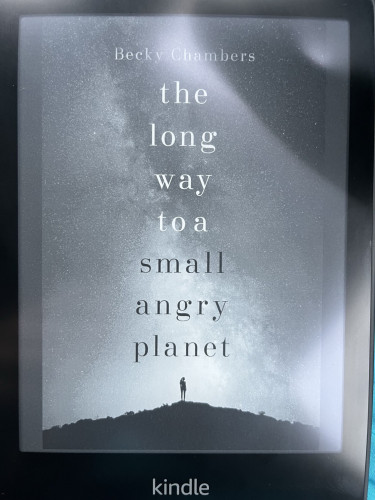 A kindle showing the front cover of ‘the long way to a small angry planet’ by Becky Chambers. It’s a simple yet effective cover, with the majority of it being covered by an immense starry sky. At the bottom is a silhouette of a lone figure standing atop a hill.