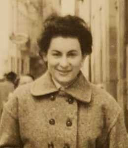 Braulia Cánovas in sepia toned B&W photo. Short dark hair swept back from faace, smiling direct at camera. Wearing wool coat with double buttoned front. Street scene in background. 