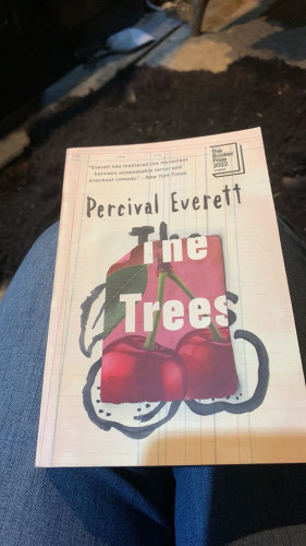 Im holding a book titled "The Trees" by Percival Everett. The cover of the book has a graphic of cherries against a lined-paper background, and a sticker for The Booker Prize 2022