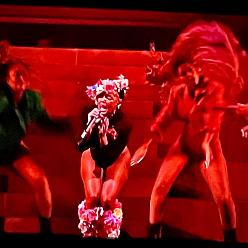 Janelle Monae in a flower crown singing, flanked by dancers tossing long braids