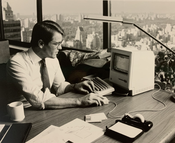 Man in office sits at desk with bank of windows and NYC cityscape in background with a bank of windows to his left. With an original Apple Macintosh computer, in 1984, sitting on the desk, his left hand on the edge of the keyboard and his right with over and moving the Mac's mouse. Coffee mug, calendar, papers, tape and a desk lamp also are in this square black and white image. The man is wearing a white dress shirt and dark tie and is looking at the Mac's small screen.