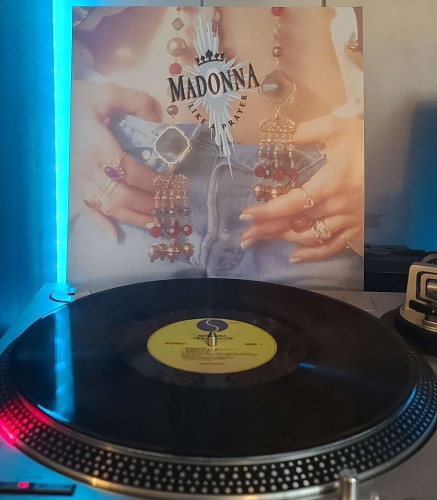 A black vinyl record sits on a turntable. Behind the turntable, a vinyl album outer sleeve is displayed. The front cover shows Madonna's bare belly and her hands resting on her jeans. Some beaded jewelry also hangs down