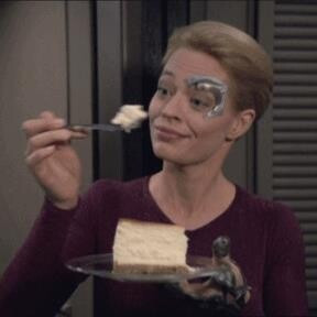 Jeri Ryan as Star Trek character Seven of Nine, holding a plate with a slice of cheesecake with one hand; with the other hand she brings a fork with a piece of the cake to her face. Her face has a very pleased expression. 
