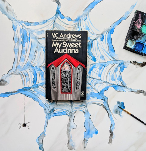 The book, My Sweet Audrina by V.C. Andrews, lying on top of a blue and black hand painted spiderweb. A spider hangs down to the left, paints are off to the right. 
