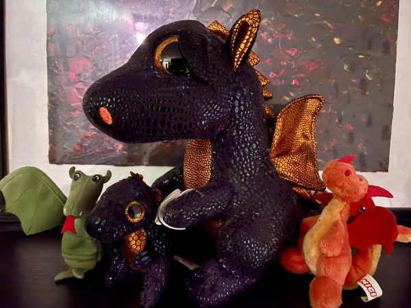 4 dragon plushies, a little green finger puppet, a little red orange dragon with a key ring, a large black and orange dragon with a little mini me between its paws. 
