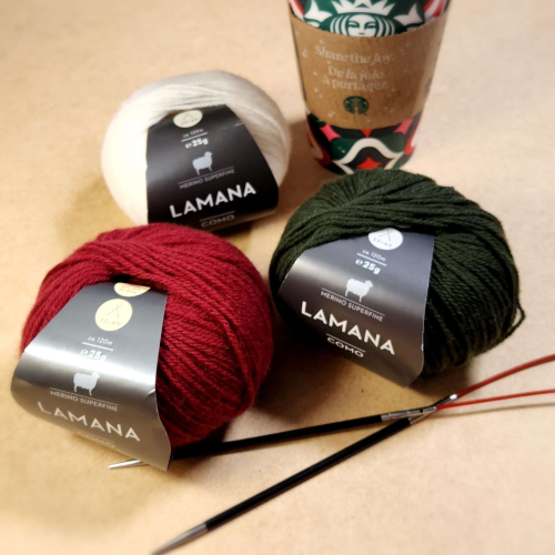 Three balls of Lamana yarn. Deep red, green and cream. A pair of cable needles sit in front and a Starbucks festive takeaway cup is in the background.