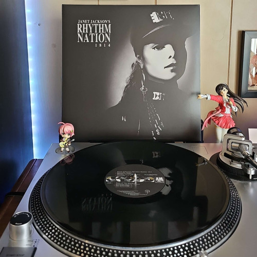 A black vinyl record sits on a turntable. Behind the turntable, a vinyl album outer sleeve is displayed. The front cover shows Janet Jackson wearing a military style cap, key earring, and a high collar military style top. 
