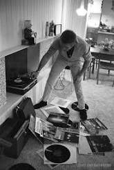 Steve McQueen bending over a collection of vinyl records scattered on the floor with a turntable nearby.