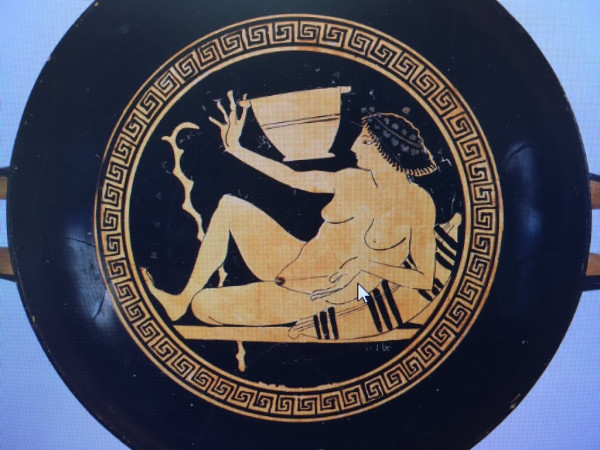 Red-figure vase painting of a female reveller playing kottabos. She is in the nude, lounging on a couch, with a walking stick in the background often shown with male revellers. She holds a cup of wine in one hand, flinging it with the other in a game of kottabos.