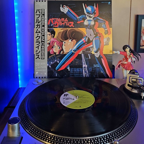 A black vinyl record sits on a turntable. Behind the turntable, a vinyl album outer sleeve is displayed. The front cover shows Priss in her Knight Saber suit, and the faces of Priss, Nene, and Sylia. 