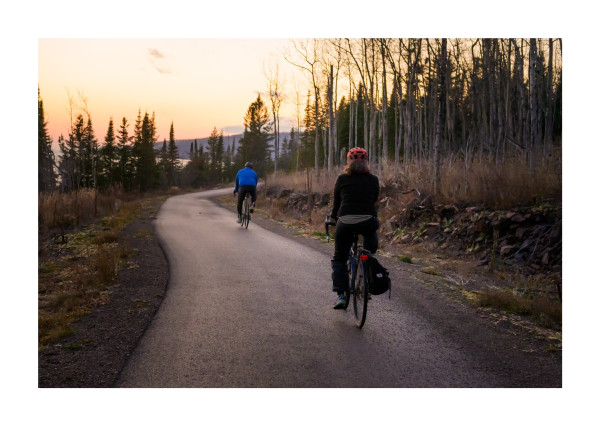 Two bikers riding on a blacktop bike path towards sunset.
