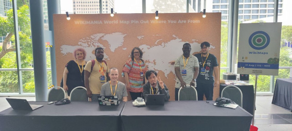 Photo of 7 people, 5 standing, 2 seated behind 2 tables and in front of a huge world map. Of to the right side is a poster board with the Wikimaps logo.