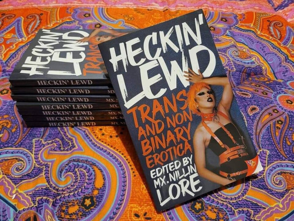 A stack of paperback copies of "Heckin' Lewd: Trans and Nonbinary Erotica" edited by Mx. Nillin Lore are stacked on top of a very colourful blanket with complex patterns.