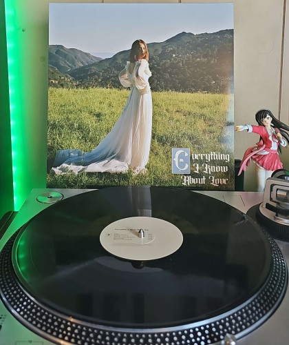 A black vinyl record sits on a turntable. Behind the turntable, a vinyl album outer sleeve is displayed. The front cover shows Laufey wearing a long whtie dress standing in a wide open field with mountains in the background. 

To the right of the album cover is an anime figure of Yuki Morikawa singing in to a microphone and holding her arm out. 