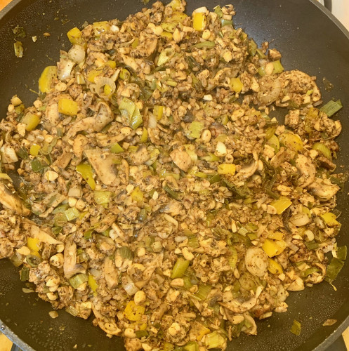 A skillet with cooked finely chopped vegetables and tempeh. Pieces of scallions, yellow-bell peper, sliced mushrooms, chopped tempeh, and rice are visible in the skillet.