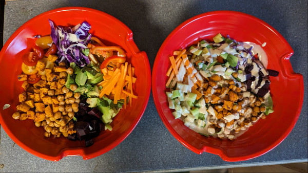 Two red bowls containing lettuce, shredded purple cabbage, carrots, red pepper, beet, sweet potato, chickpeas, cucumber, tomato, avocado and tahini dressing 