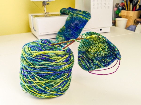 A wound cake of vibrant sock yarn in blues and greens sits on a white table. Stuck into the top of it, needles hold the beginning of a toe up sock with a simple lace pattern, almost lost among the colour changes. In the background, the completed other sock is draped very artfully indeed over my sewing machine.