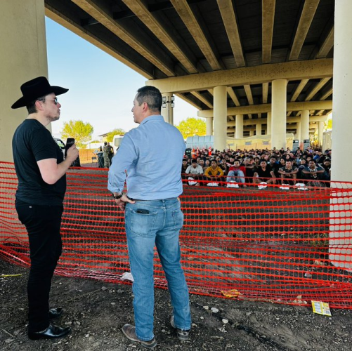 Elon Musk with a border official looking at hundreds of migrants sitting on the other side of a fence.