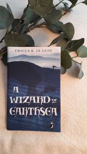 The Cover of A Wizard of Earthsea by Ursula K. Le Guin shows a landscape in blues. Far away in the back, snowed mountain ranges form a succession in light blues Closer to us, atop the line that defines the closest hill, a figure walks. The silhouette, define in dark blue, wears a cape and carries a walking stick.
The hill itself is dark blue and what at first sight appears to be grass is in fact words. The almost black shadow of a dragon, indicates ones is flying over the figure.
