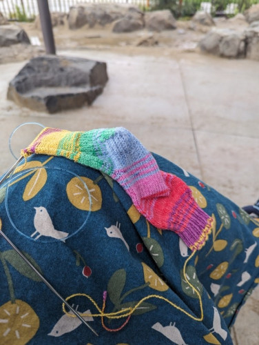 A handknit sock in progress sits on a knee covered in a handmade skirt. In the background is a damp sand/water play area at a local park.