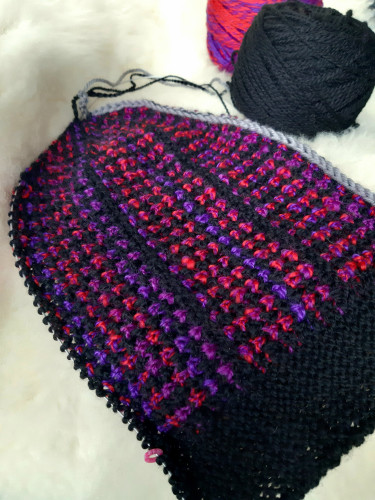 A sideways knit hat in progress. The project is knit in two yarns, one is a black and one a red and purple variegated. The brim of the bat is black, the main part in both yarns that stripe, due to tucked stitches there's a slightly dotted and textured effect. 