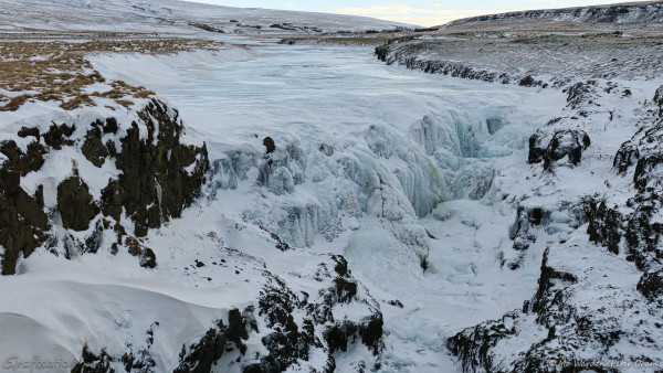 A wide river, viewed from the centre and slightly above, actually from a bridge. There are dark rocks on both banks and a waterfall has formed just in front of the viewpoint. The riversides are brown vegetation, and the whole scene is almost completely covered in snow and ice. The waterfall itself is invisible under a crust of snow-covered ice, forming strange shapes like merged icicles.