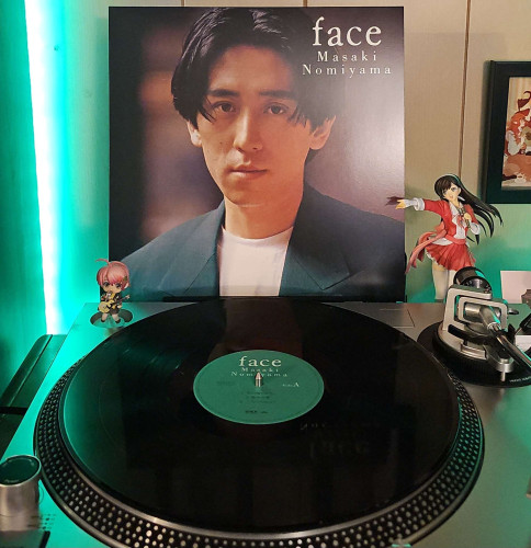 A black vinyl record sits on a turntable. Behind the turntable, a vinyl album outer sleeve is displayed. The front cover shows Masaki Nomiyama from the shoulders up. He is looking at the camera. 