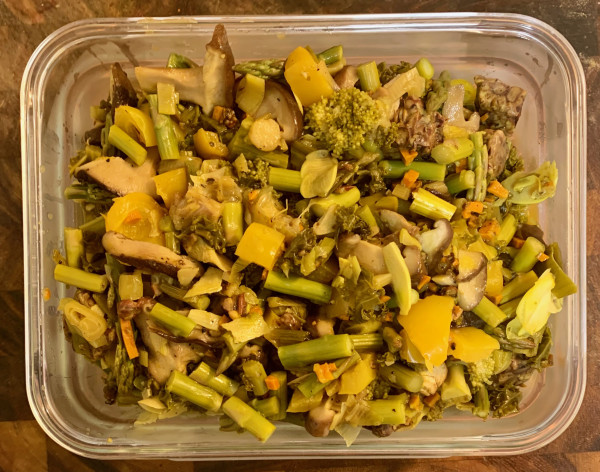 A rectangular glass storage bowl filled with a mix of cooked vegetables: asparagus, yellow bell pepper, broccoli, and kale are visible. Dish has a yellowish tint from the turmeric.