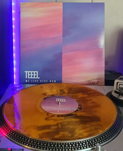 Image shows a turntable with an Orange Transparent vinyl record on the platter. Behind the turntable vinyl album outer sleeve is displayed. The front cover shows multicolored sky/clouds. The image is halved, with one side being the opposite direction of colors. Teeel's autograph is in the lower right corner