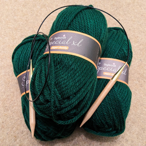 Three balls of bottle green super bulky yarn, piled up on the floor. The labels read "Stylecraft Special XL". On top of them is a pair of chonky wooden circular knitting needles.
