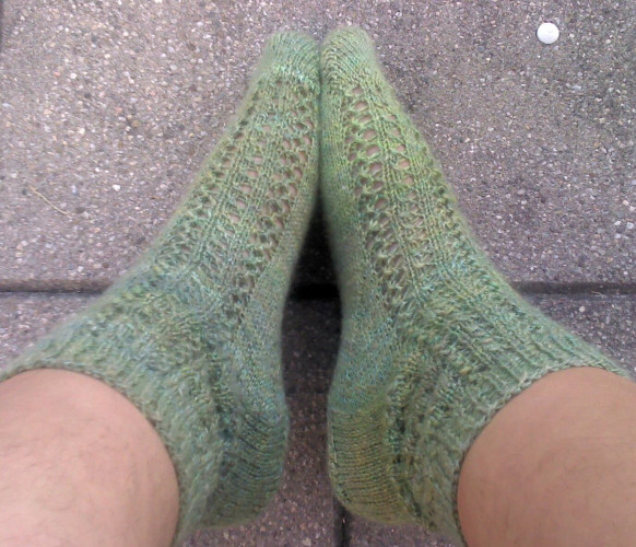 A pair of legs wearing green handknit socks with vertical lace pattern. 