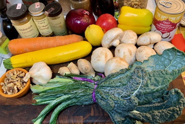 On a cutting board: 3 small jars labeled Maharajah curry, Penzey's Curry, and Marjoram, a large red onion, a small red apple, a yellow bell pepper, a can of organic coconut milk, a large Nantes carrot, a lemon, 8 large white mushrooms, a large yellow zucchini, a small bowl of walnuts, a head of garlic, 3 pieces of ginger root, and a bunch of tuscan kale.