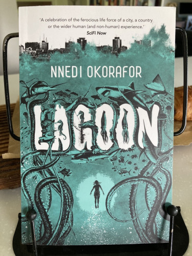 paperback: Lagoon, by Nnedi Okorafor, with a drawing of a city skyline, and sea with fish and monsters, and a person within a bright circle in the deep