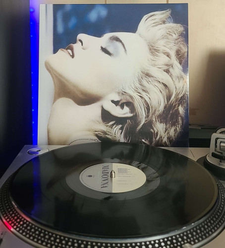 A black vinyl record sits on a turntable. Behind the turntable, a vinyl album outer sleeve is displayed. The front cover shows Madonna from the side profile. She has her head leaned back with her eyes closed. 