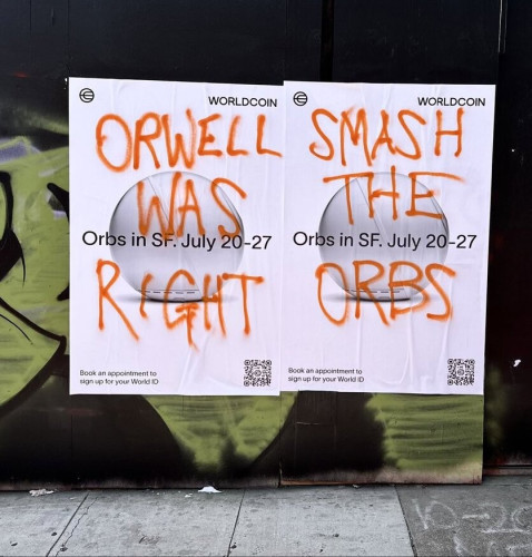Graffiti on Worldcoin posters in San Francisco, reading &quot;Orwell was right, smash the orbs&quot; in orange spray paint