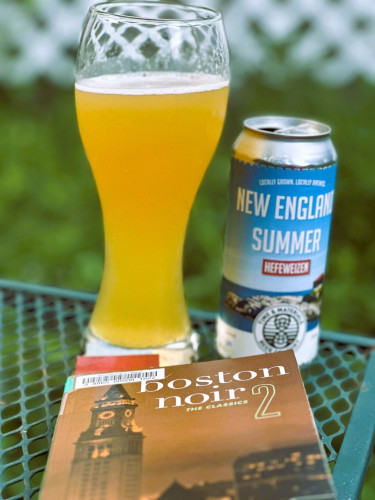 A tall wheat beer glass nearly full of a cloudy hefeweizen from Time & Materials  Beer Co, a can of which, featuring a  drawing of a rocky ocean outcropping, stands next to it. A book is in front, with a brown tinged photo of the Custom House in Boston on the cover. The book is titled boston noir The Classics.