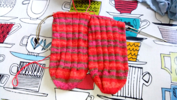 Image shows sock knitting in progress, two socks on one circular needle. Yarn is hot red and pink stripes.