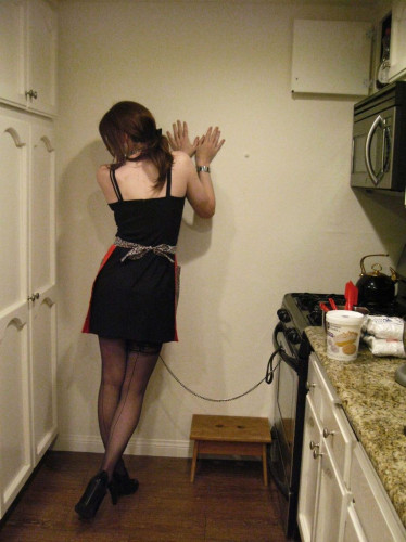 A crossdresser in a black dress has a leash attached to a stove