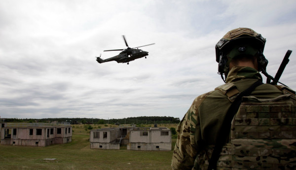 A photograph of a soldier stranding on ground while a NHIndustries NH90 multi-role military helicopter flies over a training ground made of prefab buildings.
