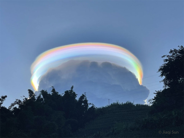 Blue sky with a dark cumulus cloud blooming up above some trees. On top of the cumulus cloud is a rainbow curving in a circle around the top, caused by light reflected on water droplets.