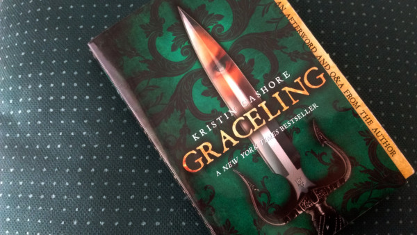 The cover illustration of Graceling by Kristin Cashore shows a a dagger, the reflection of an eye watching back at us feels the blade, resting over a green and black brocade. 