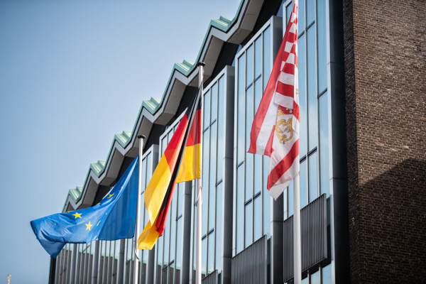 Im Hintergrund ist die Glasfront des Hauses der Bürgerschaft zu sehen. Im Vordergrund stehen drei Flaggenmasten, an ihnen wehen die EU-Flagge, die Deutschland-Flagge und die Flagge des Landes Bremen. Die Sonne scheint und der Himmel ist blau.