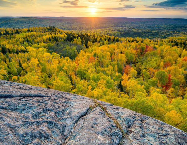 Sunset over a valley filled with maple and birch trees in full yellow and red fall color. The foreground is a rock that shows weathering from glaciers.