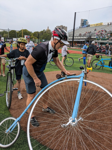 Daytime, baseball field. A man wearing a bicycle helmet lays down a pennyfarthing bicycle. In the background are four other people holding or walking with regular safety bicycles. 