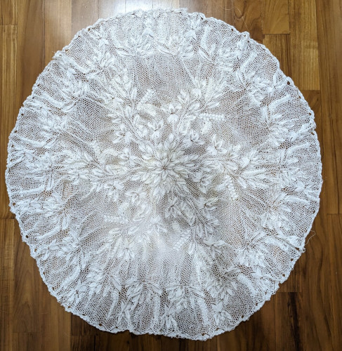 A large unblocked white lace doily, laid flat and photographed from the top. It has a winding floral and leaf motif.