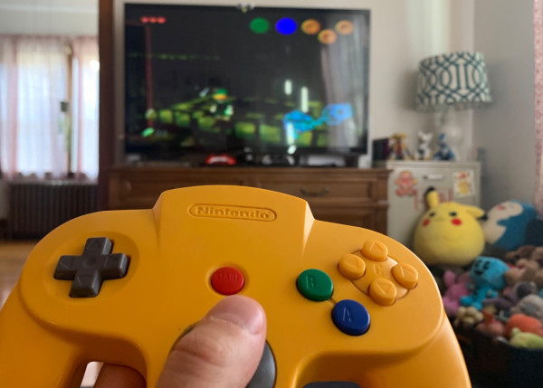 A Yellow N64 controller being held with Ocarina of Time in the background on a TV
